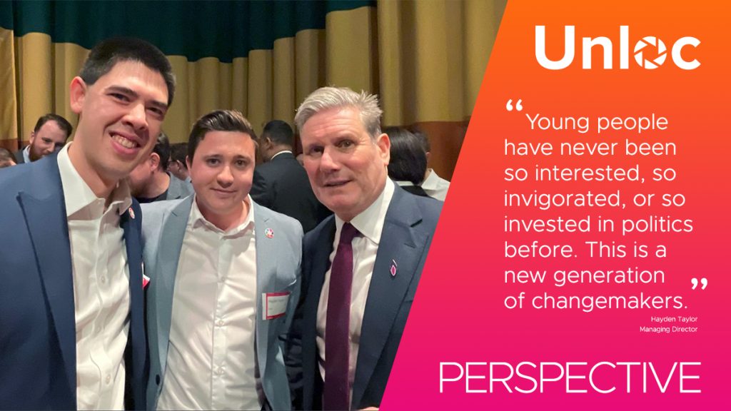 Three men in suits stand together at an event; next to them is an orange banner with a quote about young people's interest in politics and the Unloc logo.