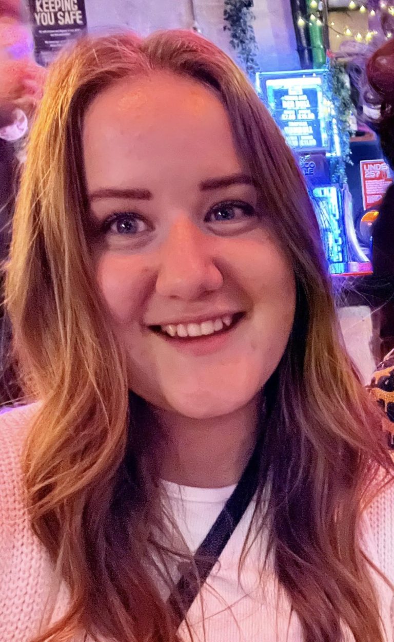 A woman with long, wavy, light brown hair is smiling at the camera in a brightly lit indoor setting with signs and neon lights in the background.