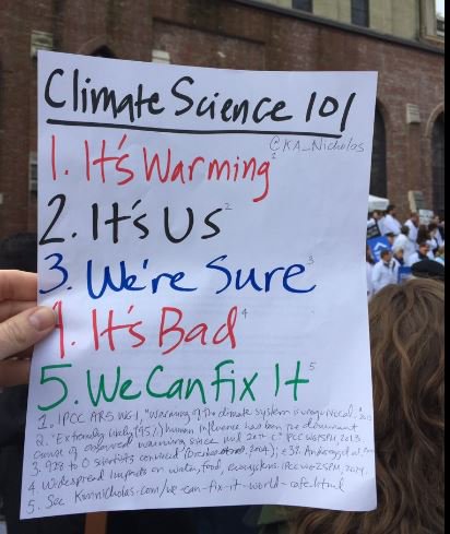A hand holds a sign titled Climate Science 101 listing five points about climate change; a crowd and building appear in the background.