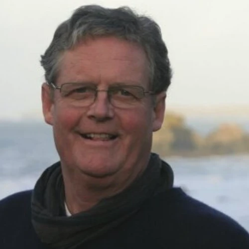 A middle-aged man with glasses and greying hair smiles at the camera with a blurred outdoor background.