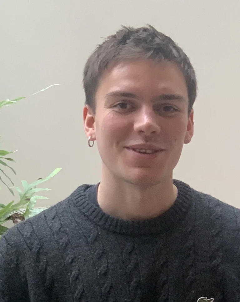 A person with short brown hair and earrings is wearing a dark cable-knit jumper, sitting in front of a light-coloured background with green plant leaves visible on the side.
