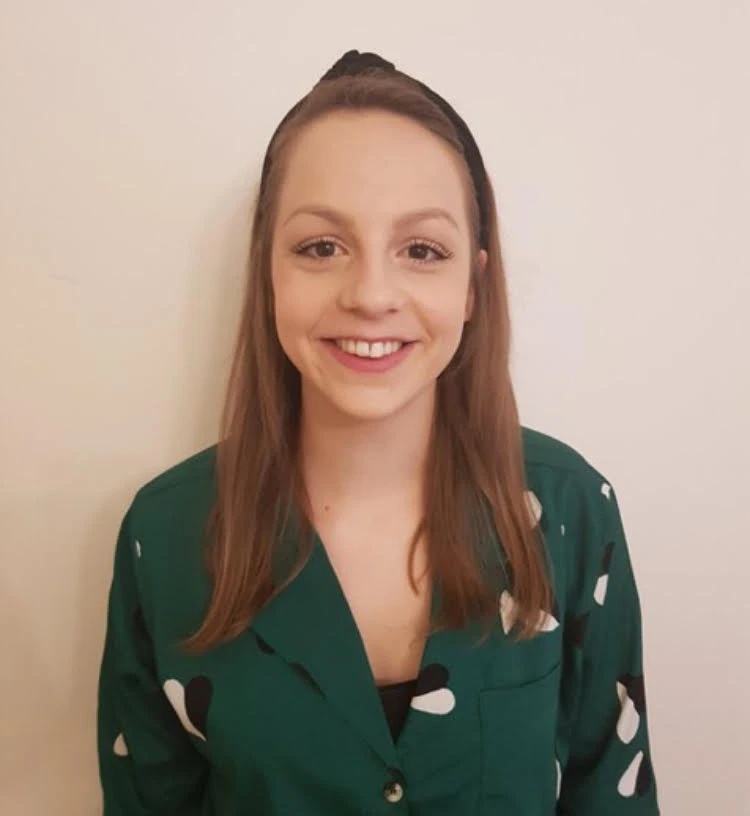 A woman with straight brown hair wearing a green patterned top and a black hairband stands in front of a plain light-coloured wall, smiling at the camera.