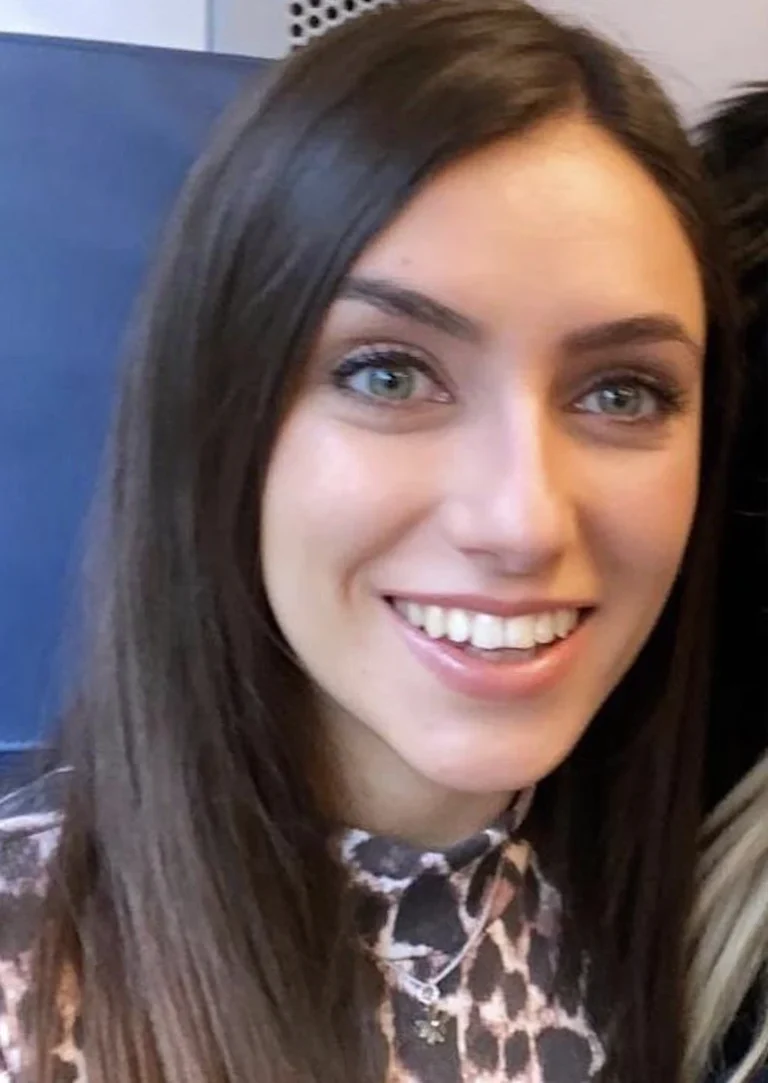 A young woman with long brown hair smiles at the camera, wearing a patterned top and a necklace, sitting indoors.
