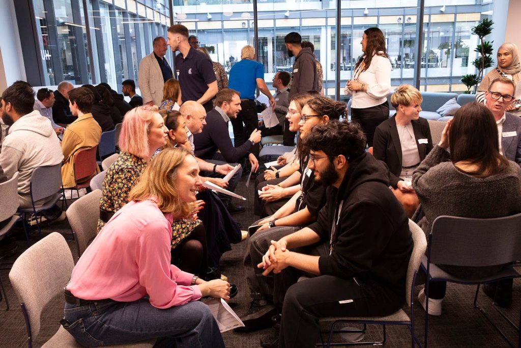 A group of people in pairs engage in face-to-face discussions in a modern office space with large windows and natural light.
