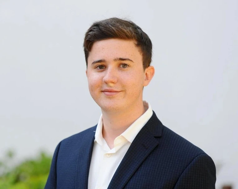 A young man with short brown hair wearing a dark suit jacket and white shirt stands in front of a plain light background.