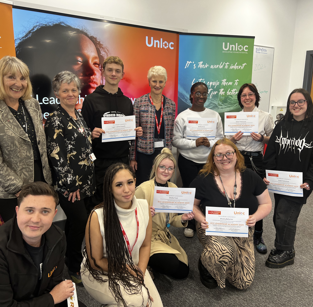 A group of people pose indoors, some holding certificates, in front of Unloc banners. Everyone is smiling and looking at the camera.