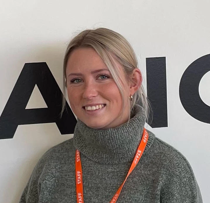 A person with blonde hair in a grey polo neck jumper and orange STAFF lanyard stands in front of a wall with large black letters.