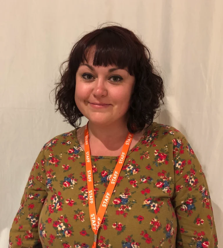 A person with dark curly hair wearing a floral shirt and an orange lanyard labelled STAFF stands against a plain light background.