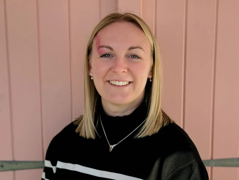 A woman with straight blonde hair and a black top stands in front of a pink wooden wall, smiling at the camera. She has a visible birthmark above her left eyebrow.
