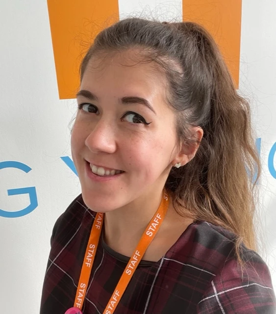 A woman with long brown hair in a ponytail, wearing a checked shirt and an orange STAFF lanyard, smiles at the camera in front of a white background with blue and orange text.