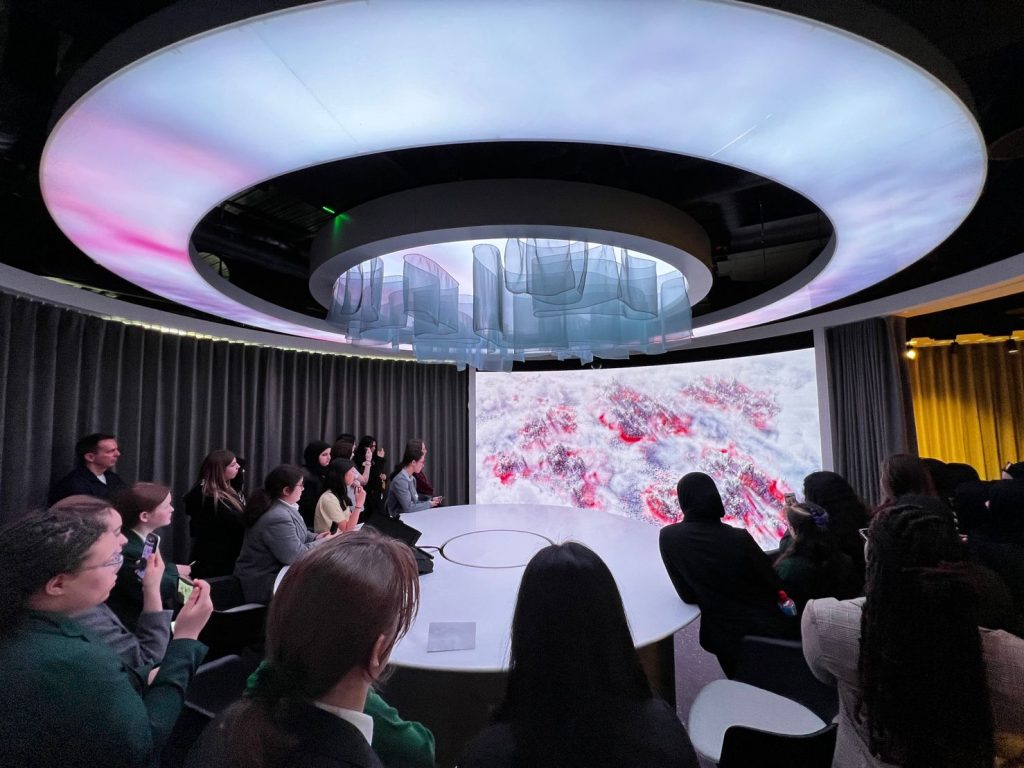 A group of people sits in a dark room around a circular table, watching a large curved screen displaying abstract red and white imagery under a futuristic ceiling.