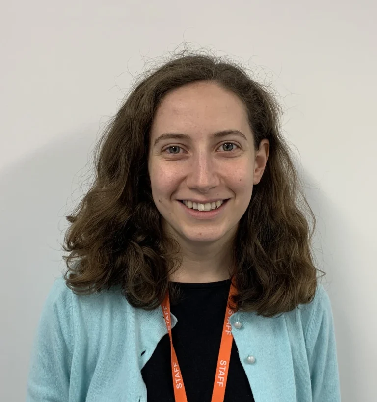 A woman with wavy brown hair wearing a blue cardigan and an orange staff lanyard stands in front of a plain white wall, smiling at the camera.