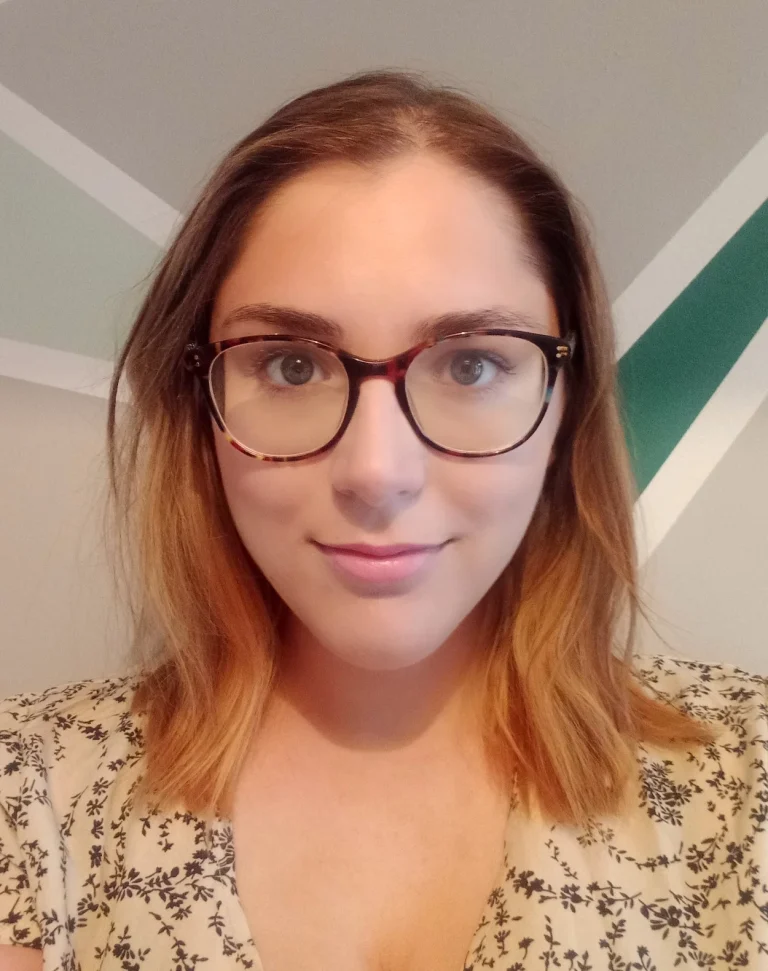 A woman with shoulder-length light brown hair and glasses looks at the camera, standing in front of a geometric-patterned wall.