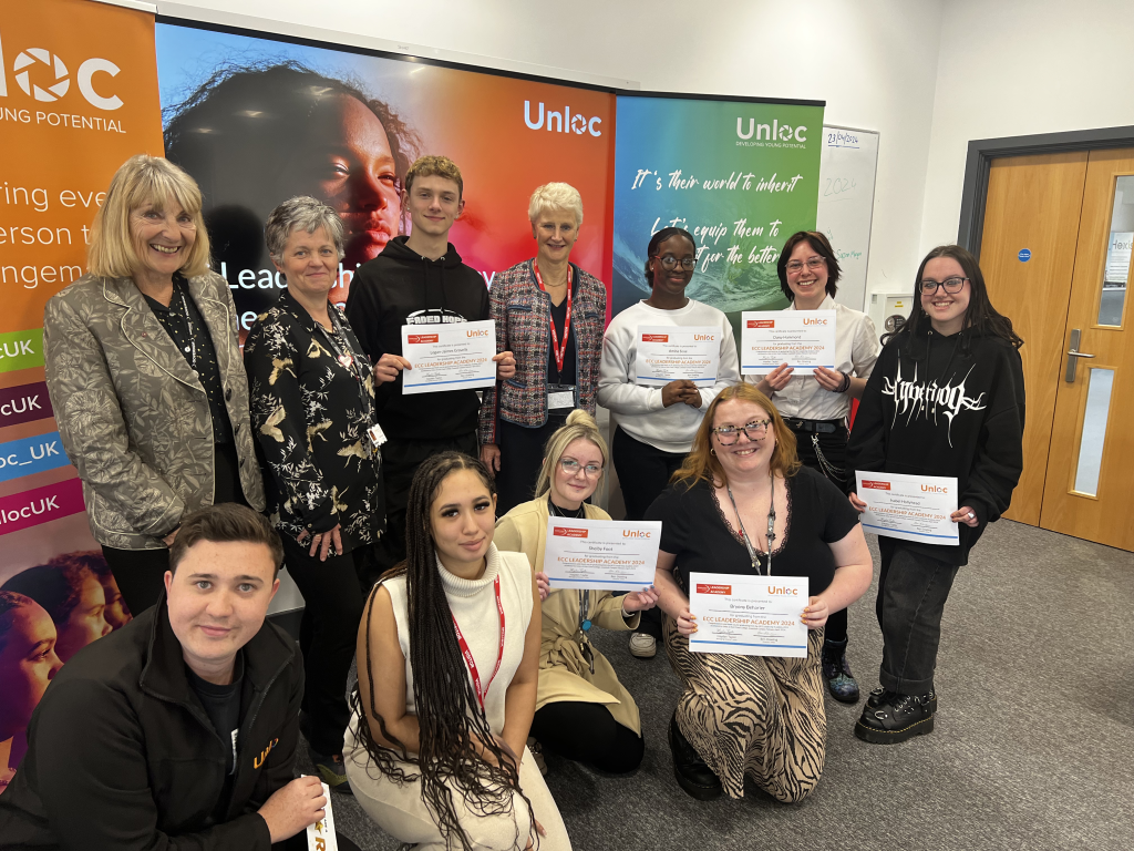 A group of people, some standing and some kneeling, pose indoors holding certificates in front of a colourful Unloc banner.
