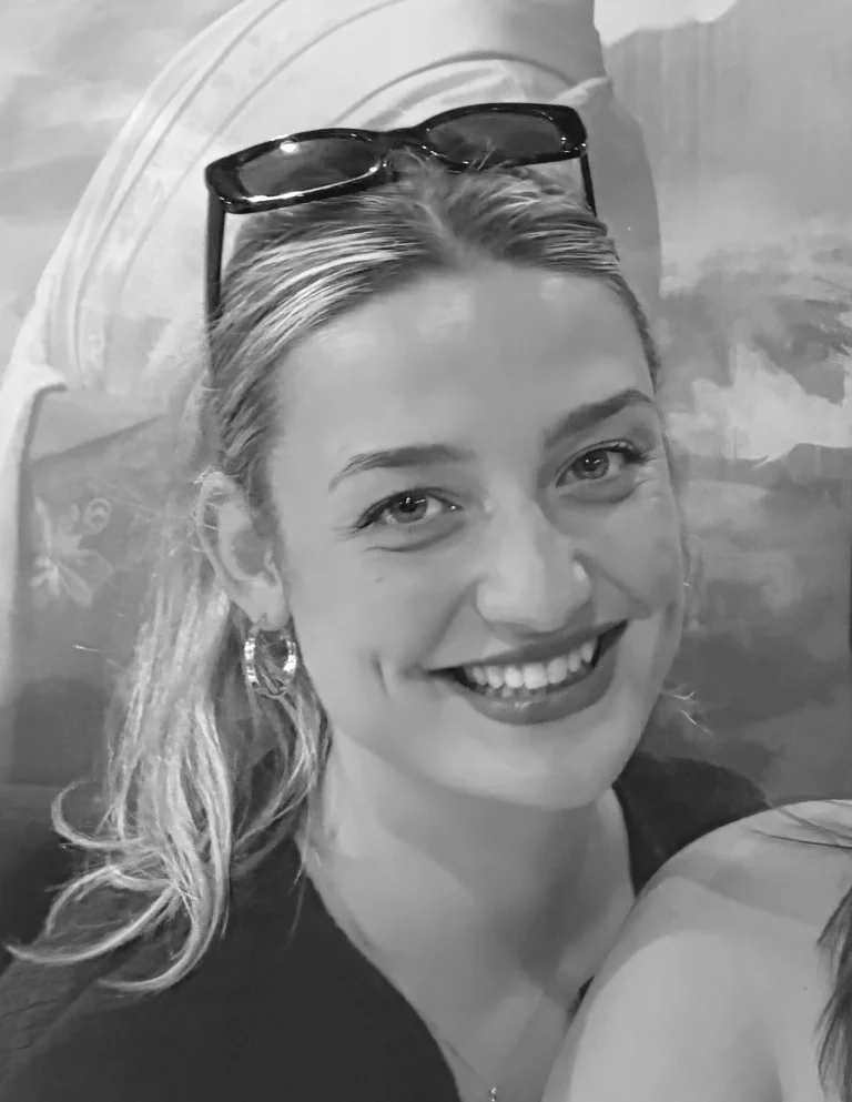 A woman with fair hair wearing sunglasses on her head and hoop earrings smiles at the camera; the photo is in black and white.