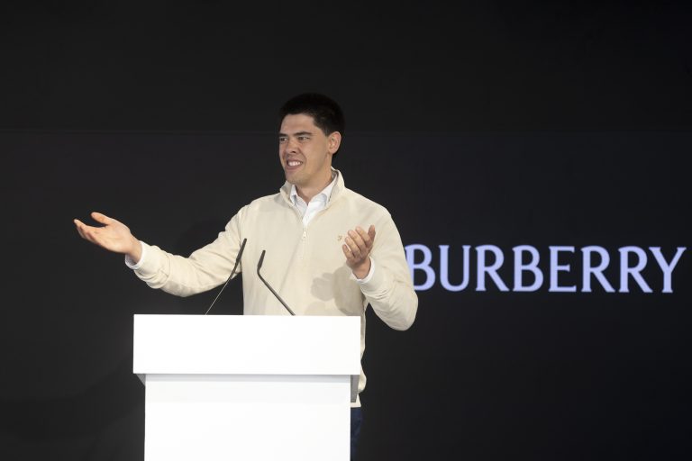 A man stands at a white lectern, gesturing with his hands, in front of a black background with the Burberry logo partially visible.