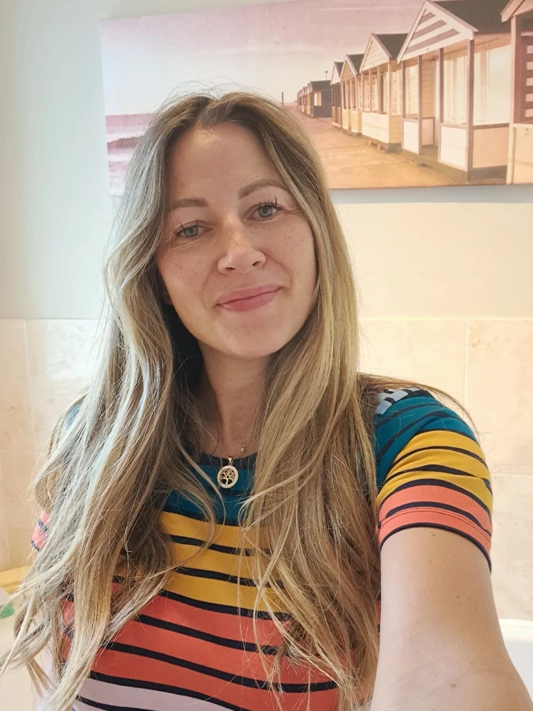 A woman with long blonde hair wearing a colourful striped top smiles at the camera; a painting of beach huts is on the wall behind her.
