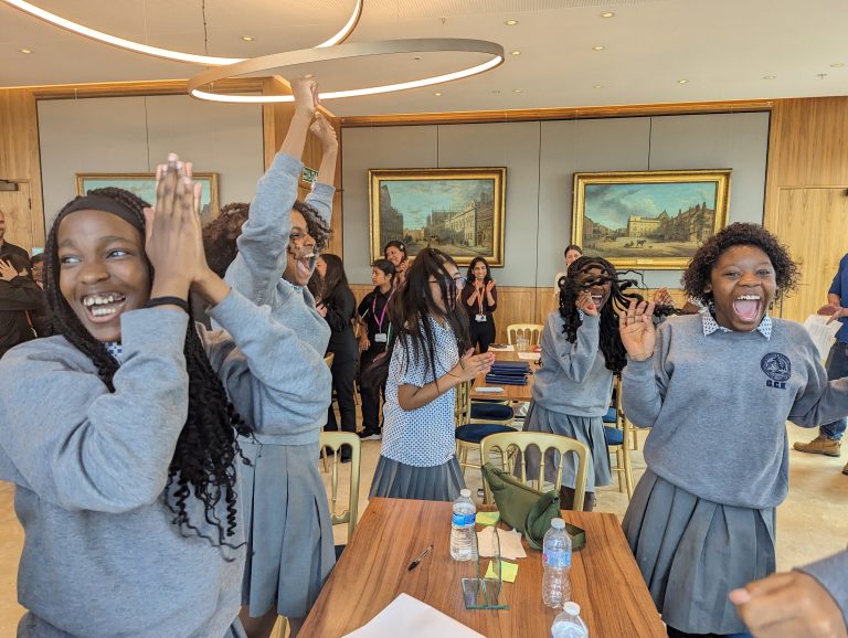 A group of students in matching grey uniforms cheer and clap around a table in a room with paintings and other people in the background.