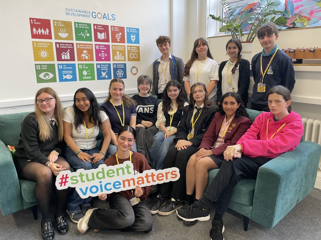 Thirteen students sit and stand in a classroom, some holding a sign reading #studentvoicematters, with sustainable development goals posters on the wall behind them.