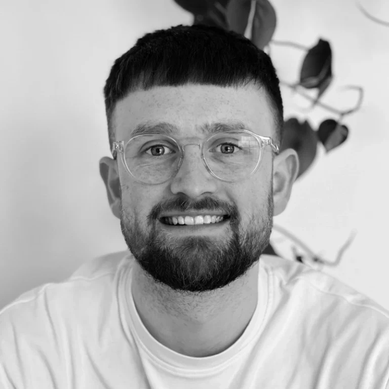 A young man with short dark hair, a beard, and clear glasses smiles at the camera. He is wearing a light-coloured shirt with a leafy plant in the background.