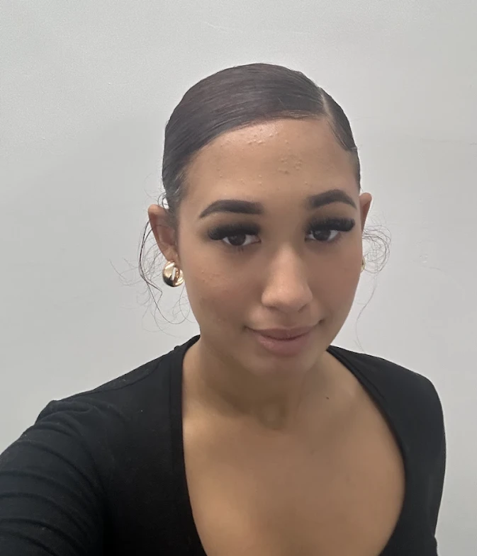 A person with dark hair pulled back, wearing gold hoop earrings and a black top, stands in front of a plain light-coloured background.