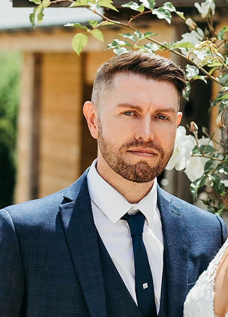 A man in a blue suit and tie stands outdoors near white flowers, looking at the camera. He has short brown hair and a trimmed beard.