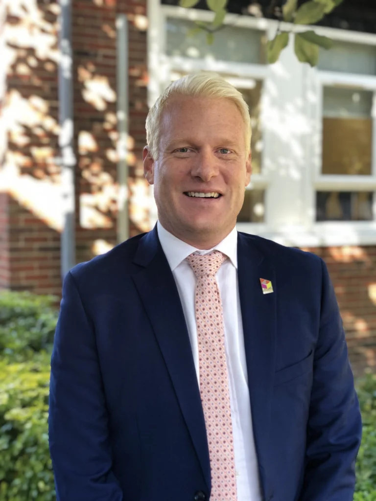 A man with blond hair in a navy suit, white shirt, and patterned pink tie stands outdoors in front of a brick building with windows.