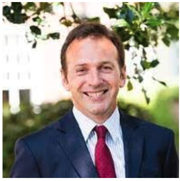 A man in a suit and red tie stands outside, smiling, with greenery and sunlight in the background.