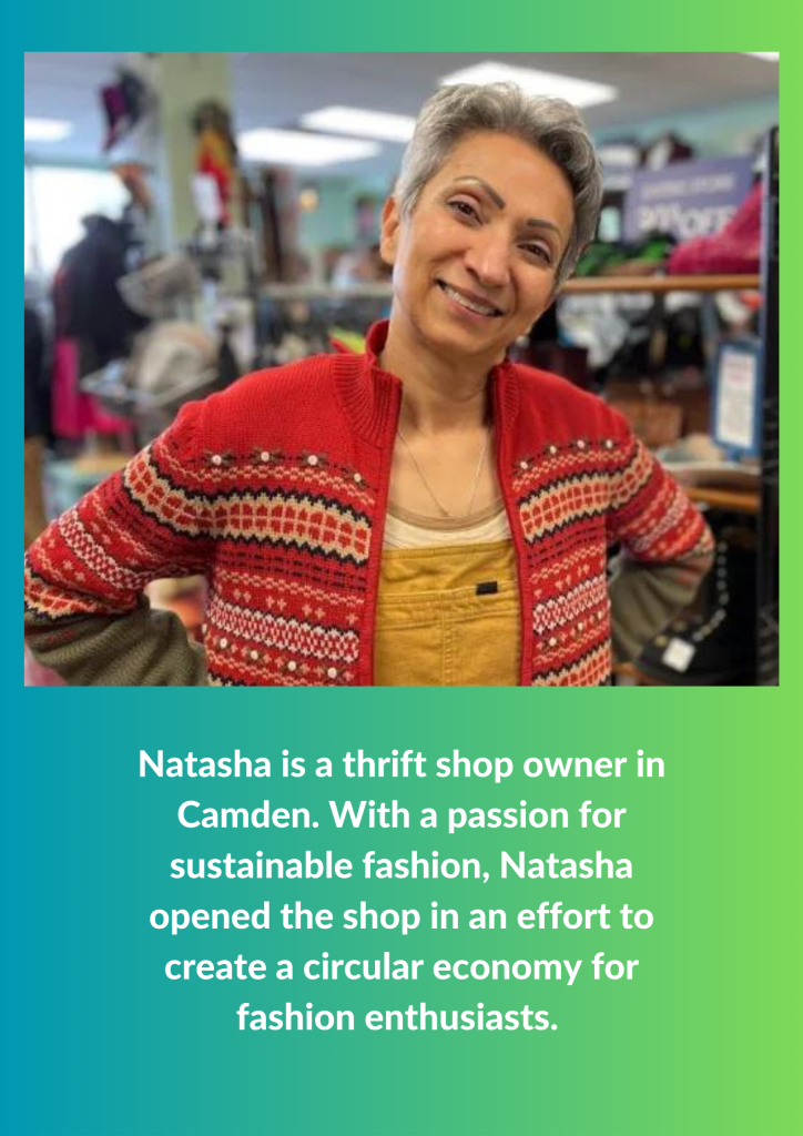 A woman wearing a red patterned jumper stands in a charity shop, smiling at the camera; second-hand clothing hangs on rails in the background.