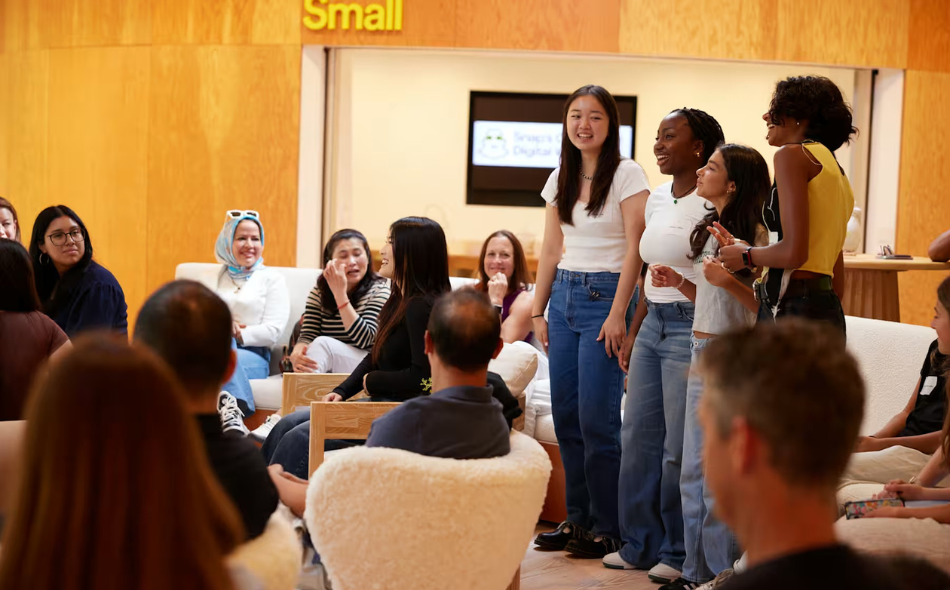 A group of young women stands and smiles whilst speaking to a seated audience in a brightly lit room with wooden walls.
