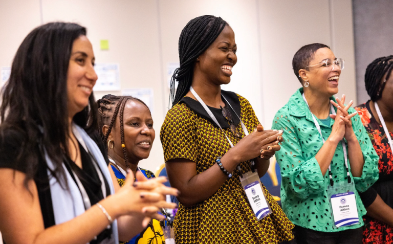 Four women stand indoors, smiling and applauding; they wear colourful clothing and conference name badges.