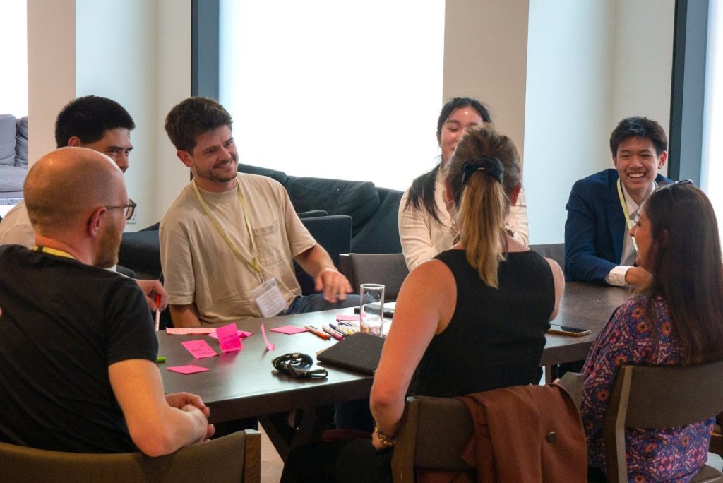 Six people sit round a table engaged in discussion, with sticky notes, pens, and notebooks in front of them.