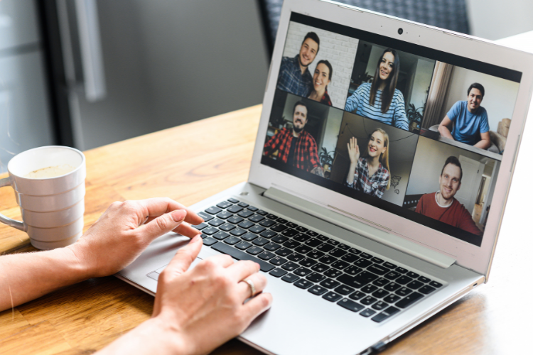 Person using a laptop for a video call with six participants, each visible in individual windows on the screen. A cup of coffee sits on the wooden table nearby.