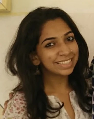 A young woman with long dark hair smiles at the camera, wearing a light-coloured patterned top, standing indoors against a light background.