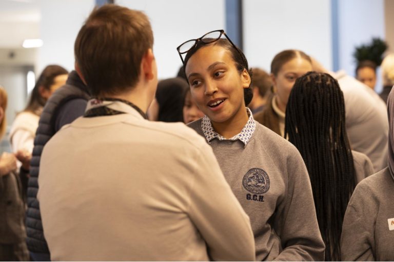 A group of people wearing grey uniforms engage in conversation at an indoor event. One young woman faces a man, speaking and smiling.