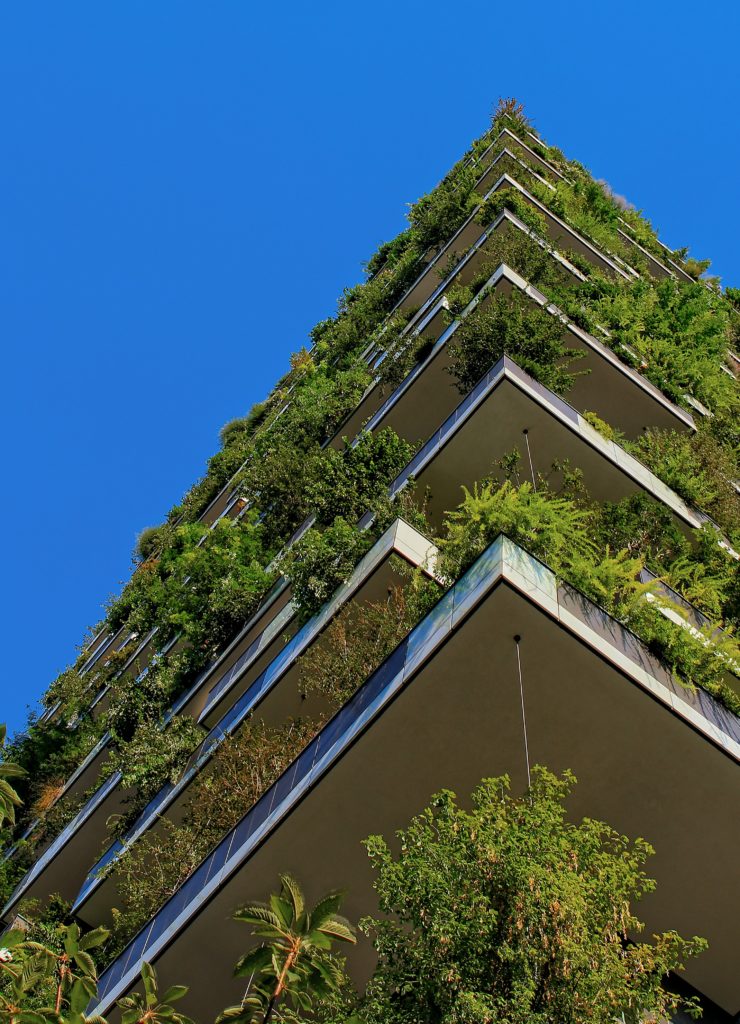 A modern building with multiple balconies covered in green plants, viewed from below against a clear blue sky.