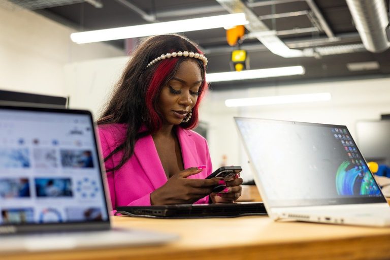 A woman in a pink blazer sits at a desk using her phone, with two open laptops in front of her, preparing materials for a training programme in a modern office setting.