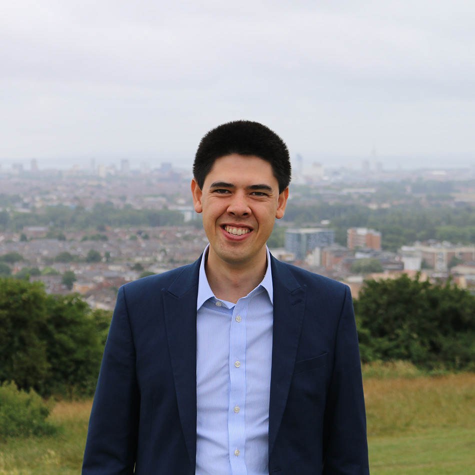 A man in a blue suit and light blue shirt stands outdoors on grass with a cityscape and cloudy sky in the background.