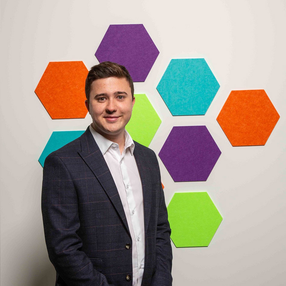 A man in a suit stands in front of a wall decorated with colourful hexagon shapes arranged in a cluster.