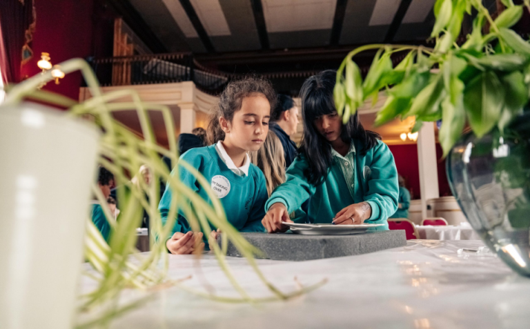 Two girls in turquoise uniforms focus on an activity at a table, surrounded by green plants, in a large, elegant indoor setting.