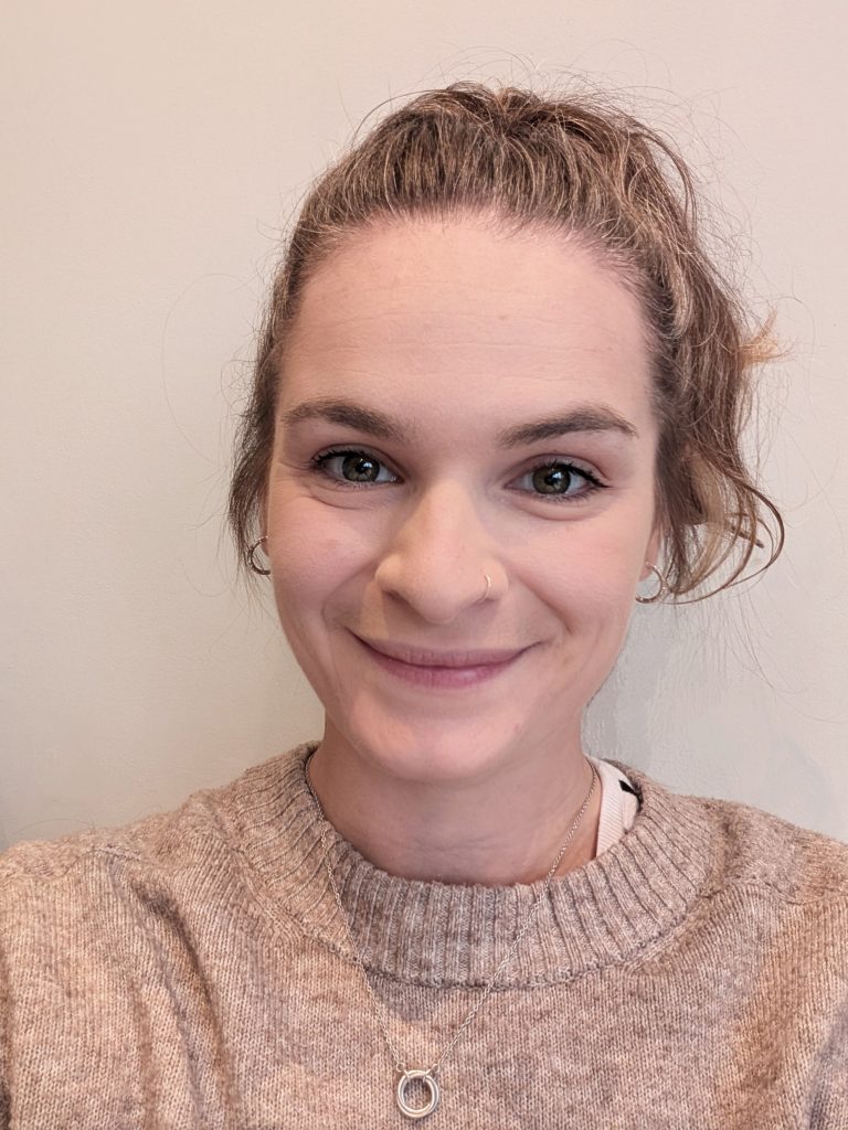 A person with light-brown hair tied back into a ponytail smiling at the camera.
