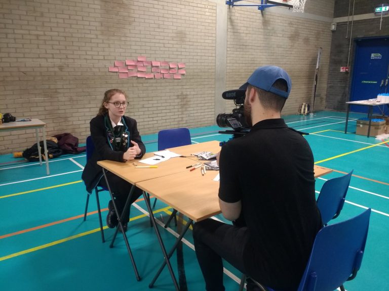 A man with a camera films a female student sitting at a table in a gymnasium, with stationery items and pink sticky notes on the wall behind her.