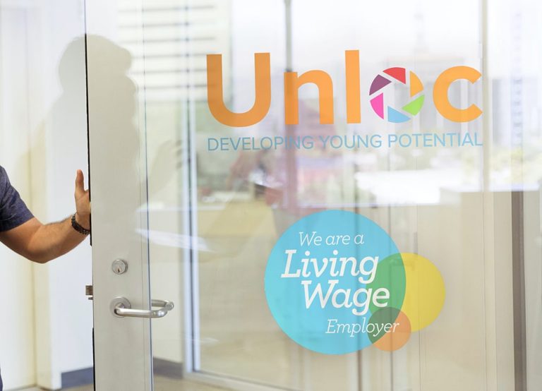 A person opens a glass door with the Unloc logo, the phrase Developing Young Potential, and a blue We are a Living Wage Employer sticker visible.