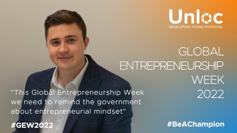 A young man in a suit sits in front of a promotional backdrop for Unloc and Global Entrepreneurship Week 2022 with a quote about reminding the government of the entrepreneurial mindset.