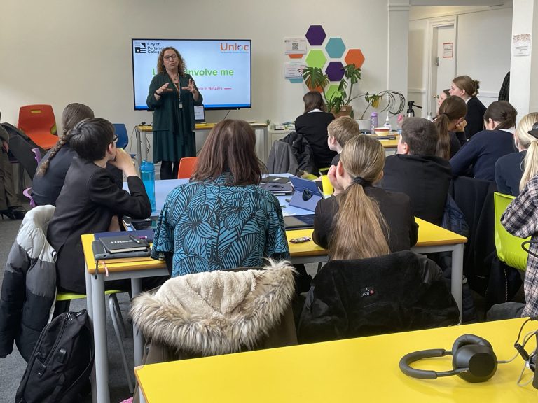 A woman stands in front of a classroom giving a presentation to students seated at desks, with a screen displaying Unlock and Involve Me as part of a training programme.