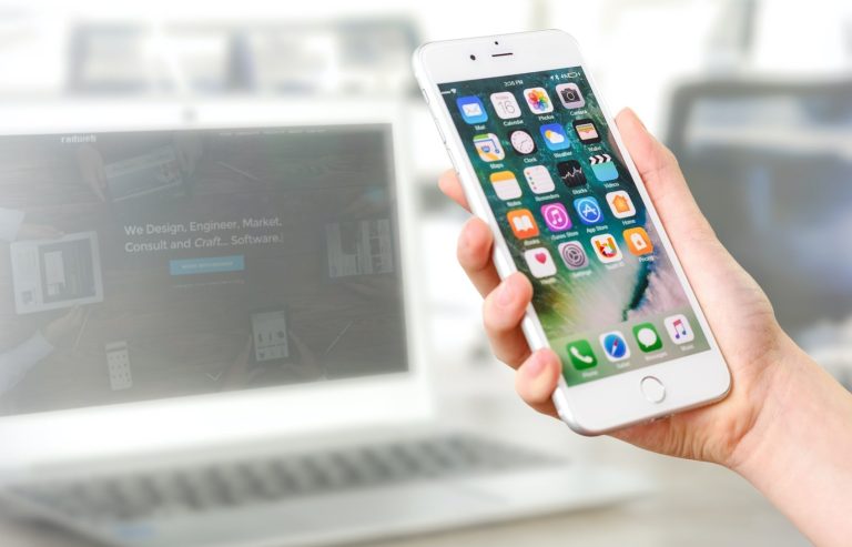 A person holds a white mobile phone displaying various app icons, with an open laptop blurred in the background.