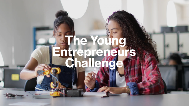 Two young women work on a small robotics project at a table in a classroom. Overlaid text reads: “The Young Entrepreneurs’ Challenge”.