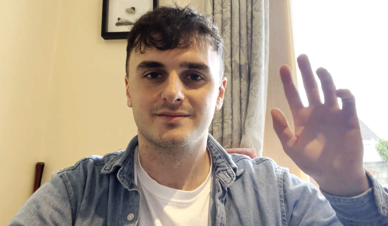 A person with short brown hair wearing a white shirt and denim jacket is sitting indoors, looking at the camera and waving with their right hand.