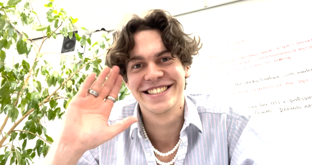 A person with curly hair, wearing a striped shirt, rings, and a beaded necklace, smiles and waves at the camera in a bright room with plants and a whiteboard in the background.