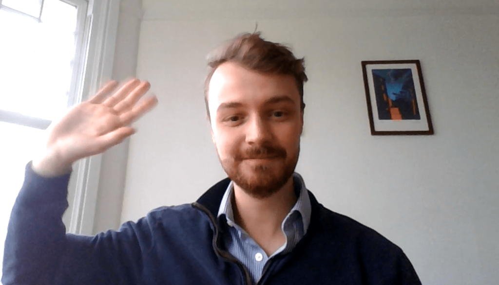 A man with light brown hair and a beard waves at the camera, sitting indoors near a window with a framed picture on the wall behind him.